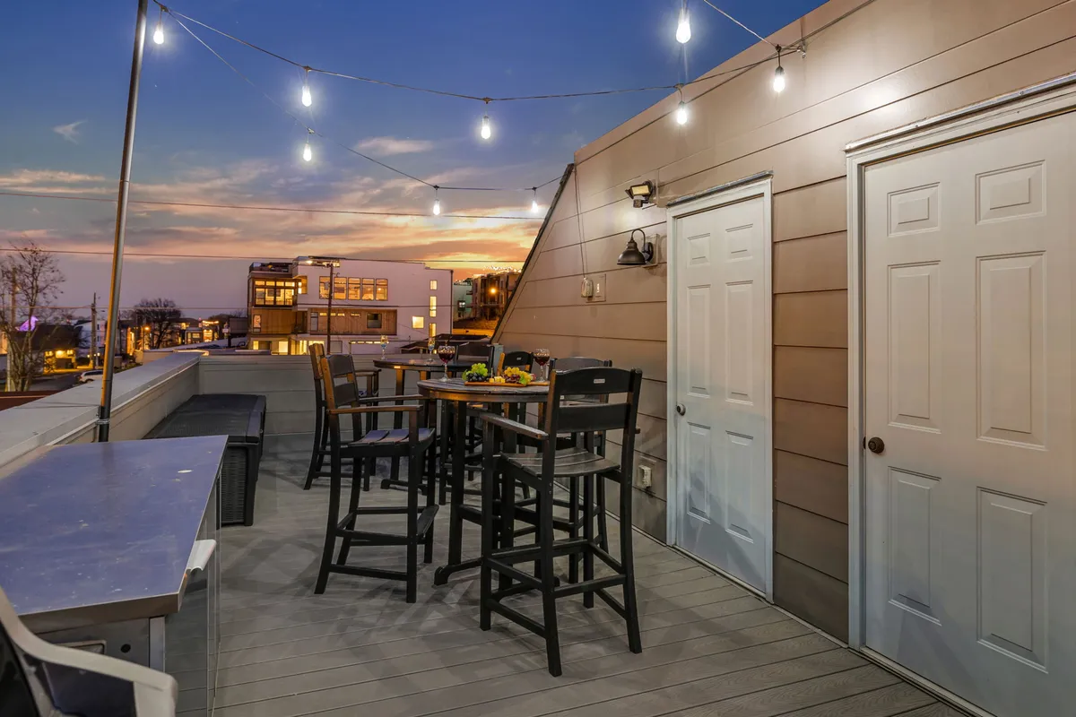 An exterior shot of an outdoor seating area illuminated by string lights with a gorgeous sunset sky in the background. The space is set for evening gatherings, with multiple tables and a clear view of the city, evoking a sense of celebration and relaxation.