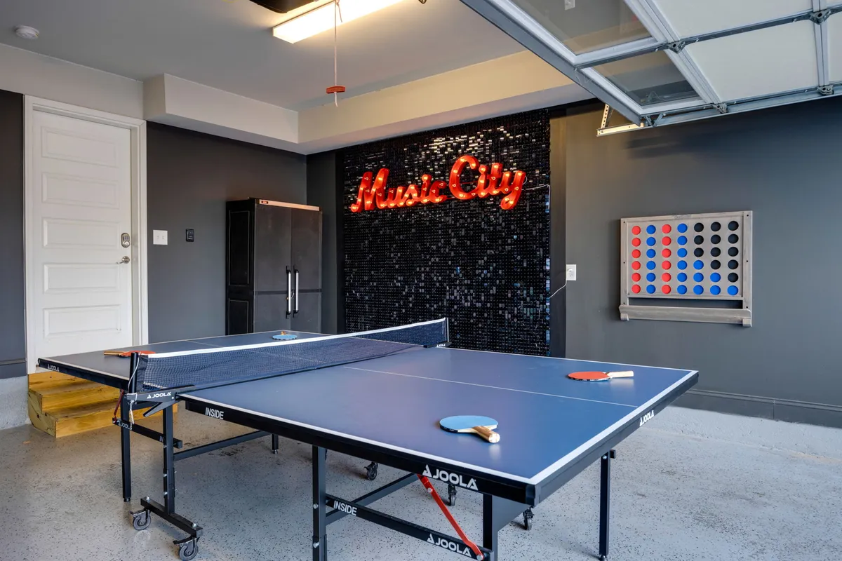 A lively interior shot featuring a ping pong table prominently in the converted garage game room. The space is energized by a neon sign, and the wall displays a colorful game board, creating a playful and inviting atmosphere perfect for group activities and game nights.