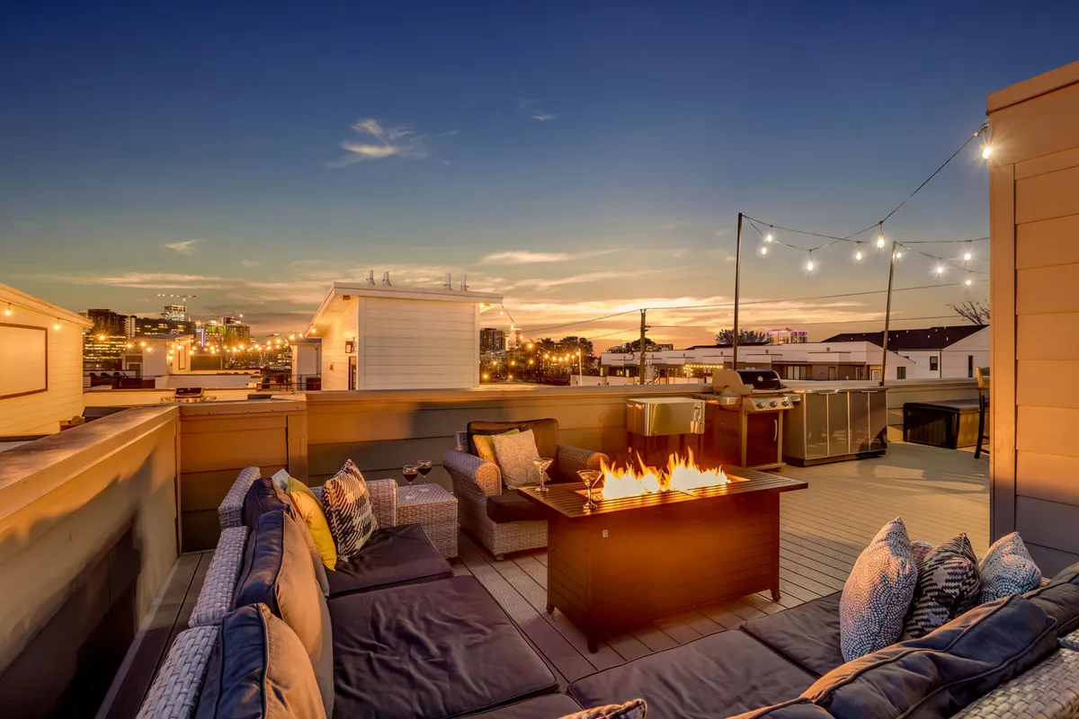 A cozy rooftop setup with comfortable seating surrounding a fire pit, string lights overhead, and a direct view of the city skyline. The inviting arrangement is ideal for socializing, dining al fresco, or relaxing under the stars.