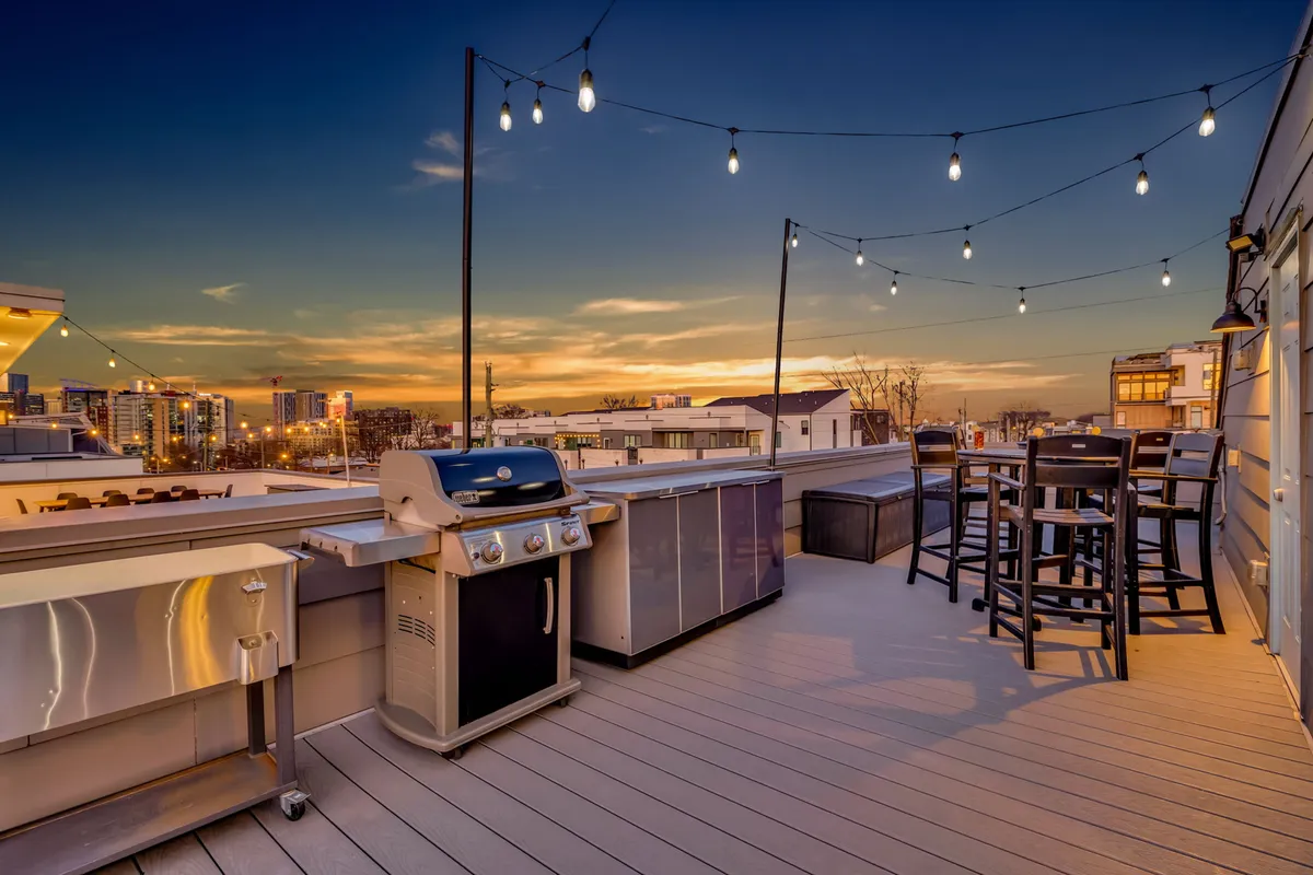 An open-air rooftop with a modern grill, bar-height seating, string lights, and a panoramic skyline view. The space is set up for barbecuing, outdoor dining, and casual drinks as the sun goes down over Nashville.