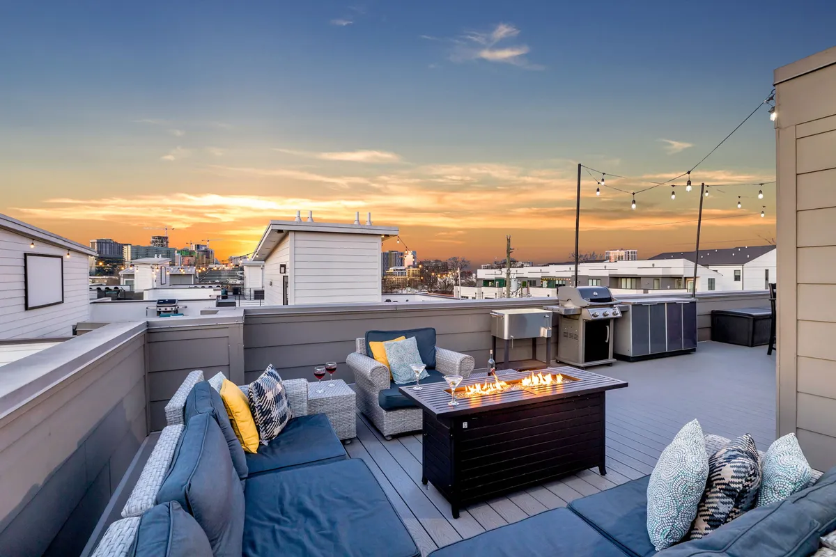 This image captures a rooftop lounge area at sunset, featuring a cozy fire pit, modern outdoor seating, a BBQ grill station, and sweeping views of the Nashville city skyline. String lights add a festive touch, and the space is designed for groups to relax and socialize while enjoying the cityscape.