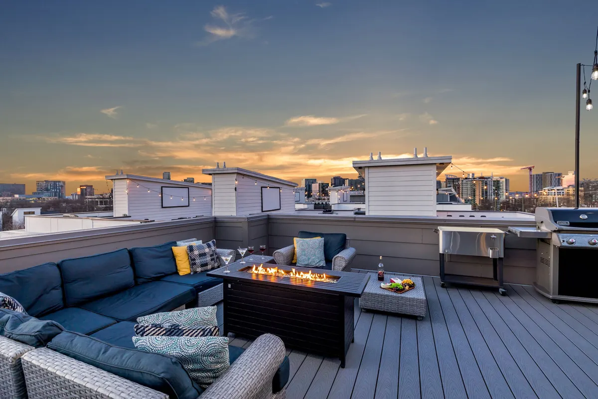 A stunning private rooftop deck featuring a welcoming fire pit encircled by modern outdoor seating, a grill station for barbecuing, and the sweeping Nashville skyline as a backdrop. String lights add warmth to the scene as dusk settles in.