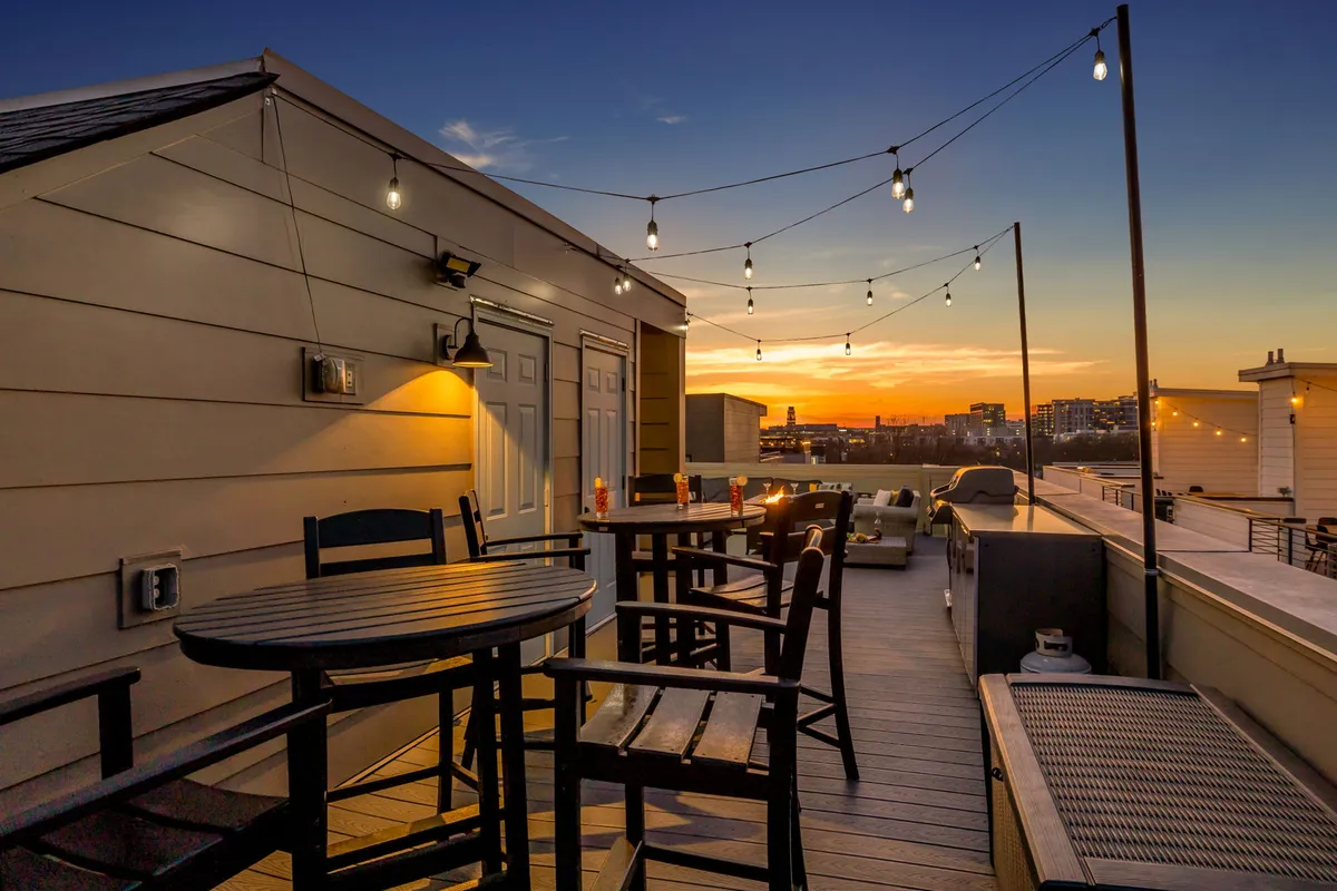 An inviting rooftop or terrace exterior shot with a wooden table, bar stools, string lights, and a beautiful sunset view. The relaxed yet celebratory atmosphere invites groups to gather for evening drinks or late-night laughs.